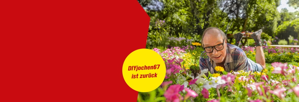 Ein Mensch mit Brille liegt lachend in einem bunten Blumenbeet.